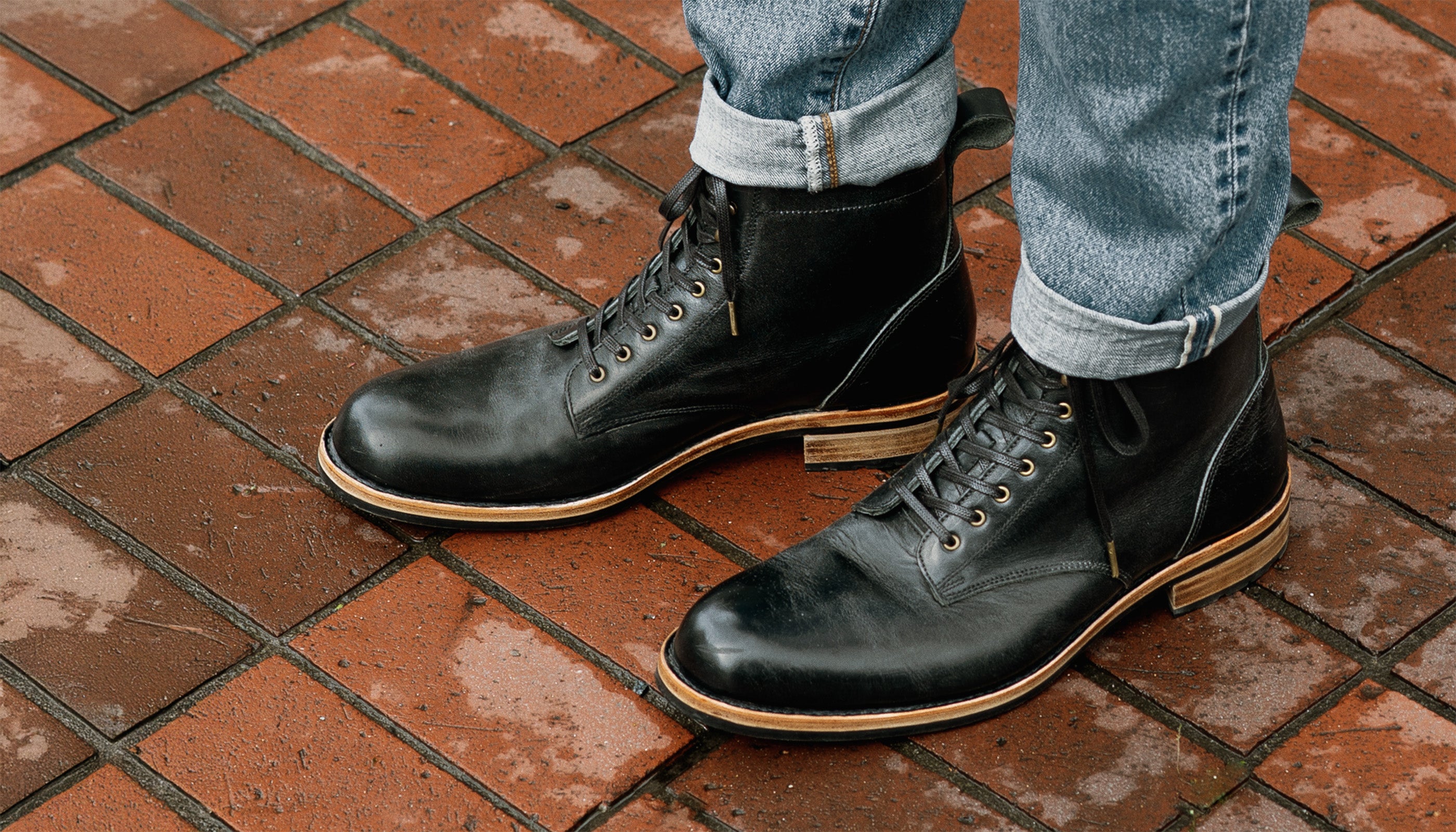 Black leather boots worn with jeans on a brick sidewalk.