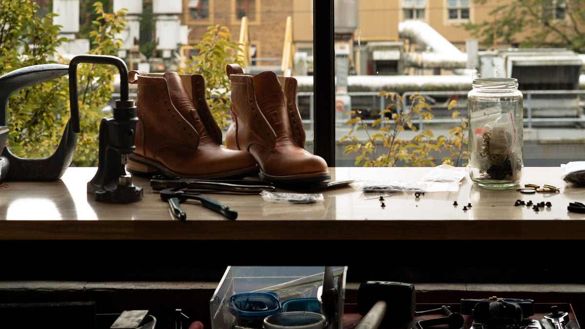 Boots in progress on a workbench in front of a window overlooking the city.