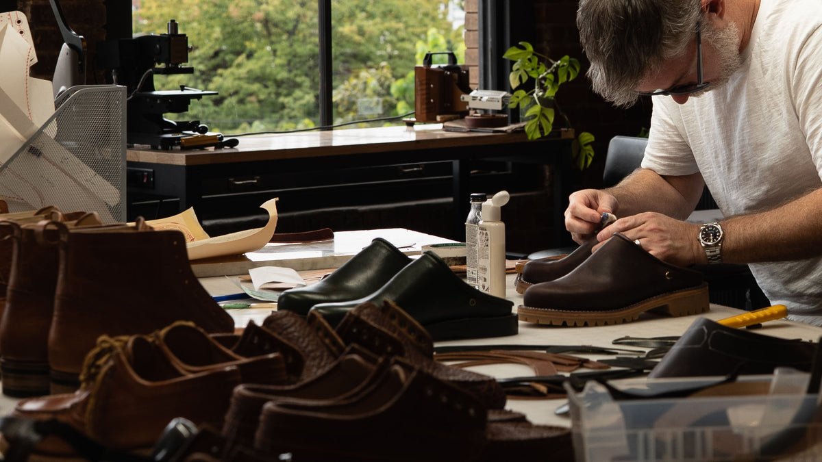 A maker at the bench in the Archive workshop, with boots in progress on the table.